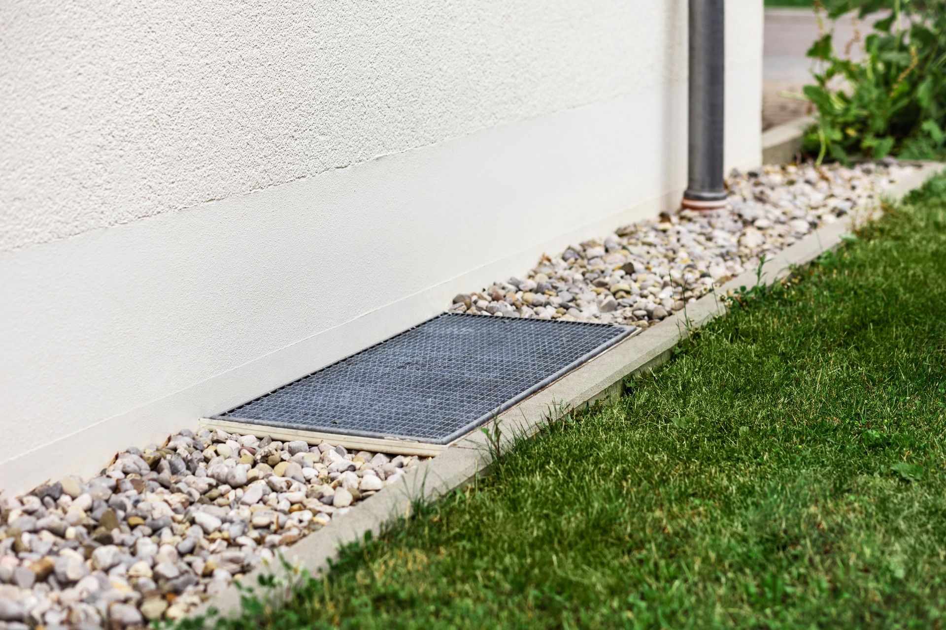 Concrete foundation with gravel and a metal grate next to a white wall and green grass.