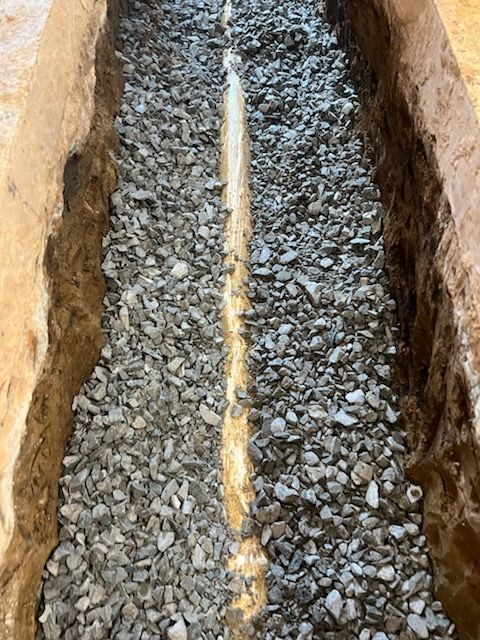 Gravel-filled trench with a central yellow pipe running down the length. Brown soil borders the trench.