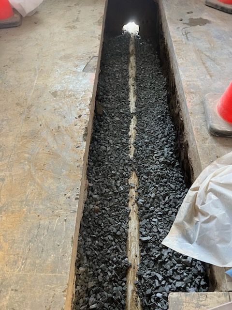 Trench filled with gravel, centered with a pipe. Concrete floor with orange cones.