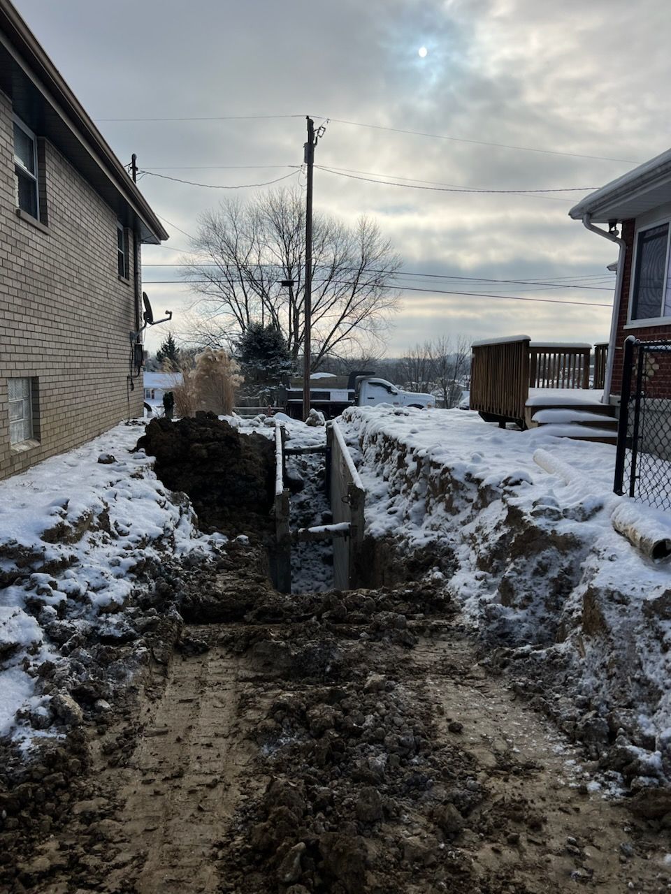 Trench dug in snowy yard between two houses. Metal supports in trench. Cloudy sky overhead.