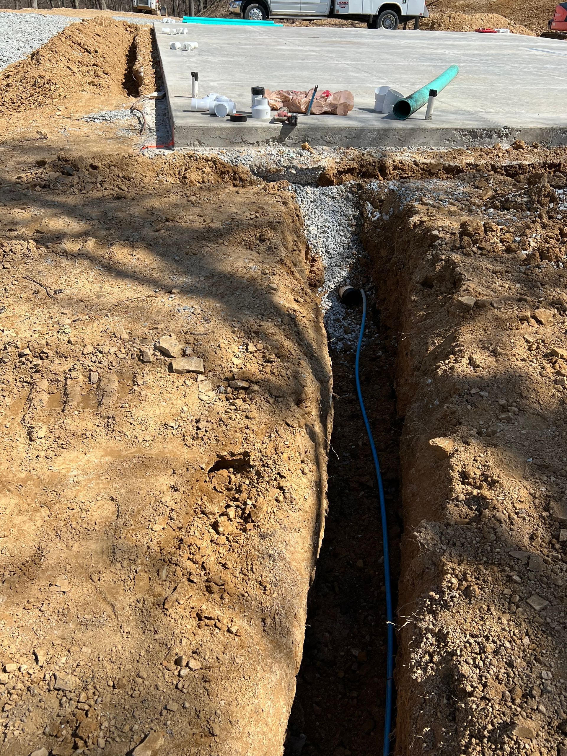Trench with blue utility line, gravel bed, and tools, likely for construction on dirt and concrete.