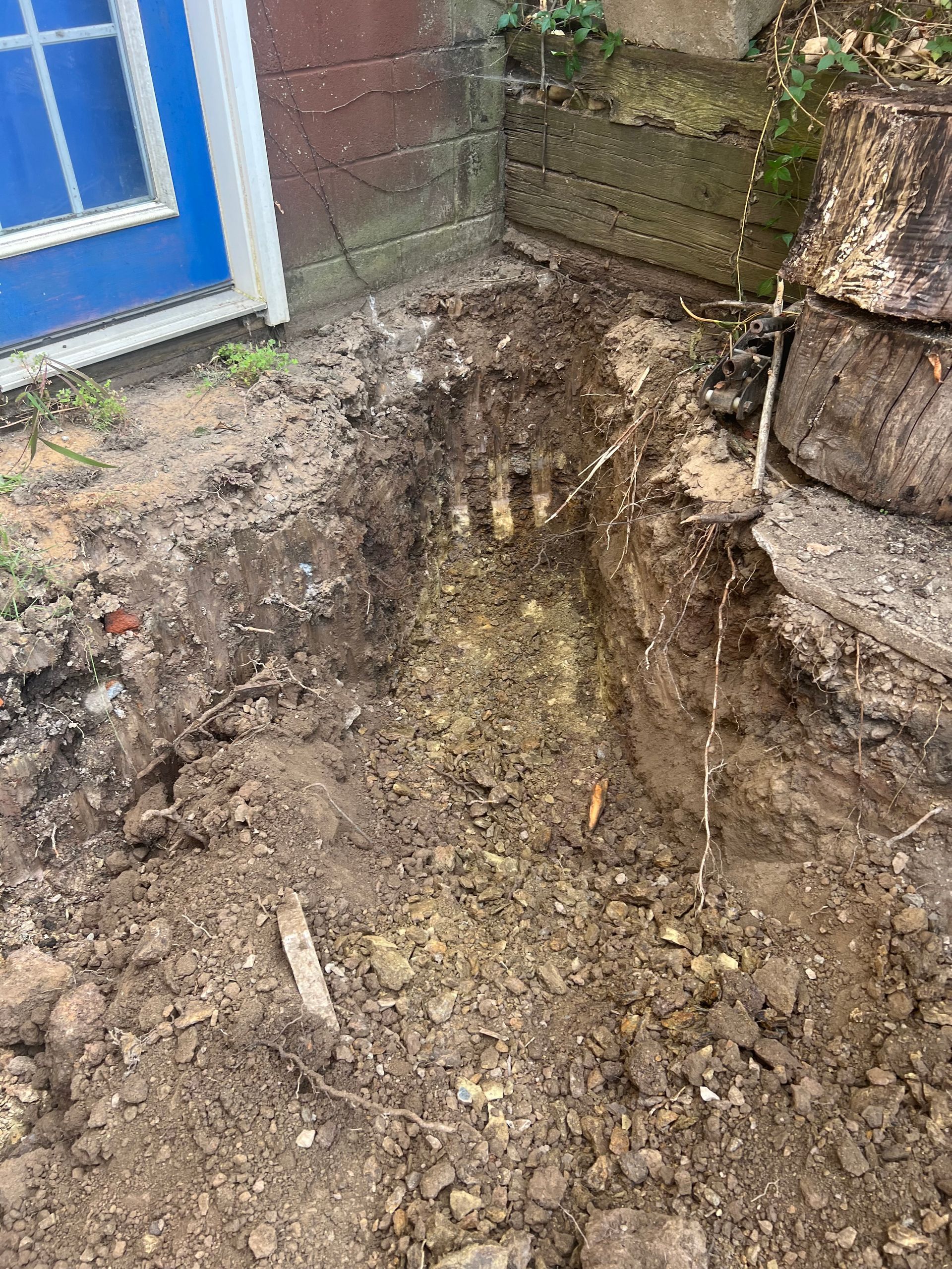 Hole dug near a blue door and brick wall, revealing three pipe ends.