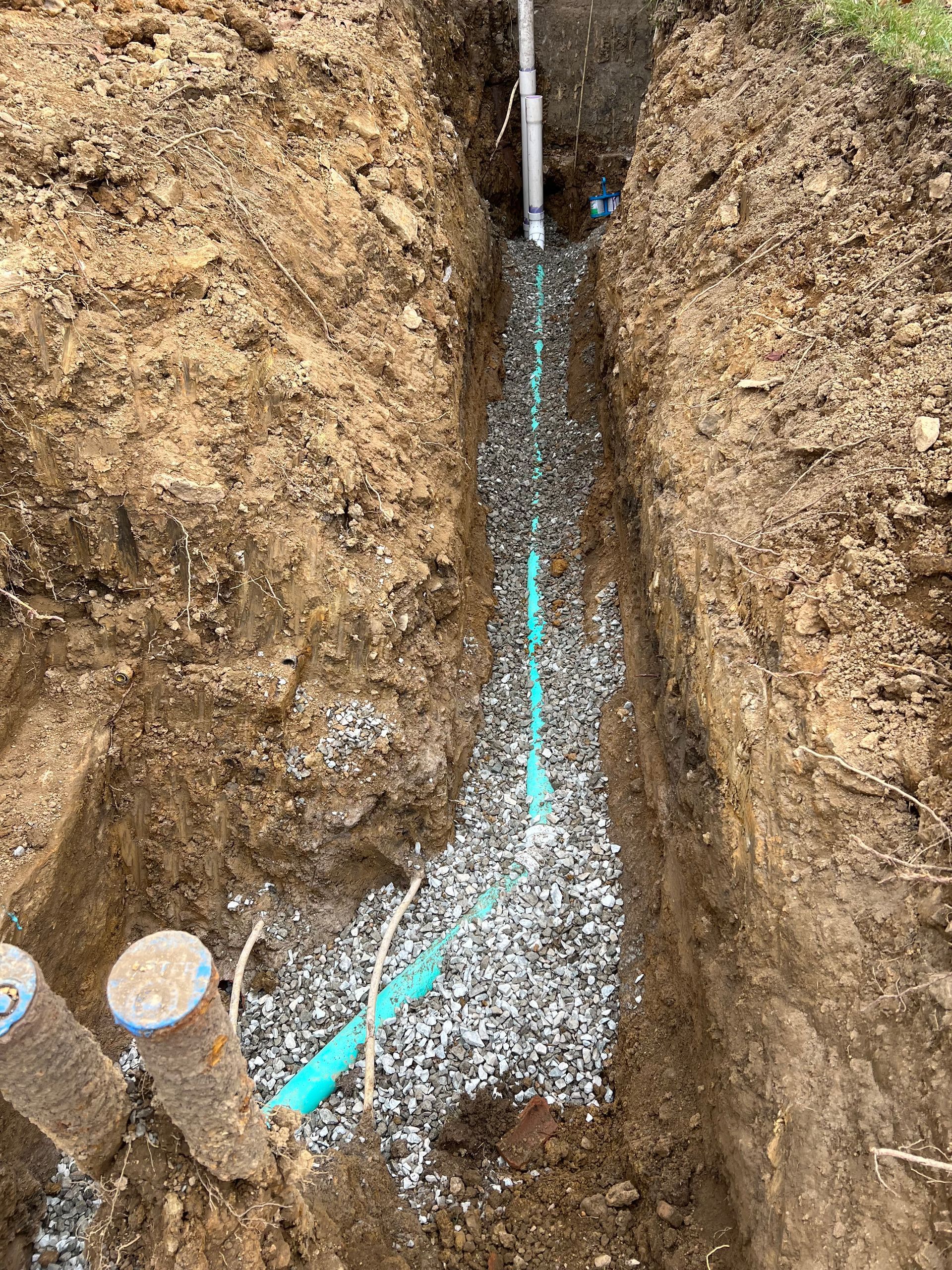 Trench with green pipe laid in gravel, possibly for water supply. Vertical metal pipe and stubs on the left.