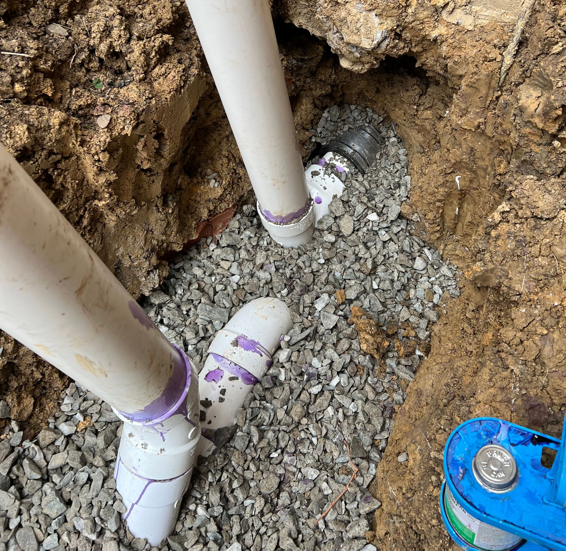 Pipes in a gravel-filled trench. PVC pipes are white with purple primer. A blue tool is visible.