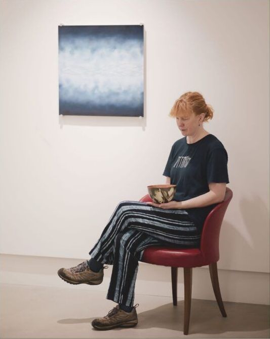 A woman sits in a chair holding a bowl in front of a painting