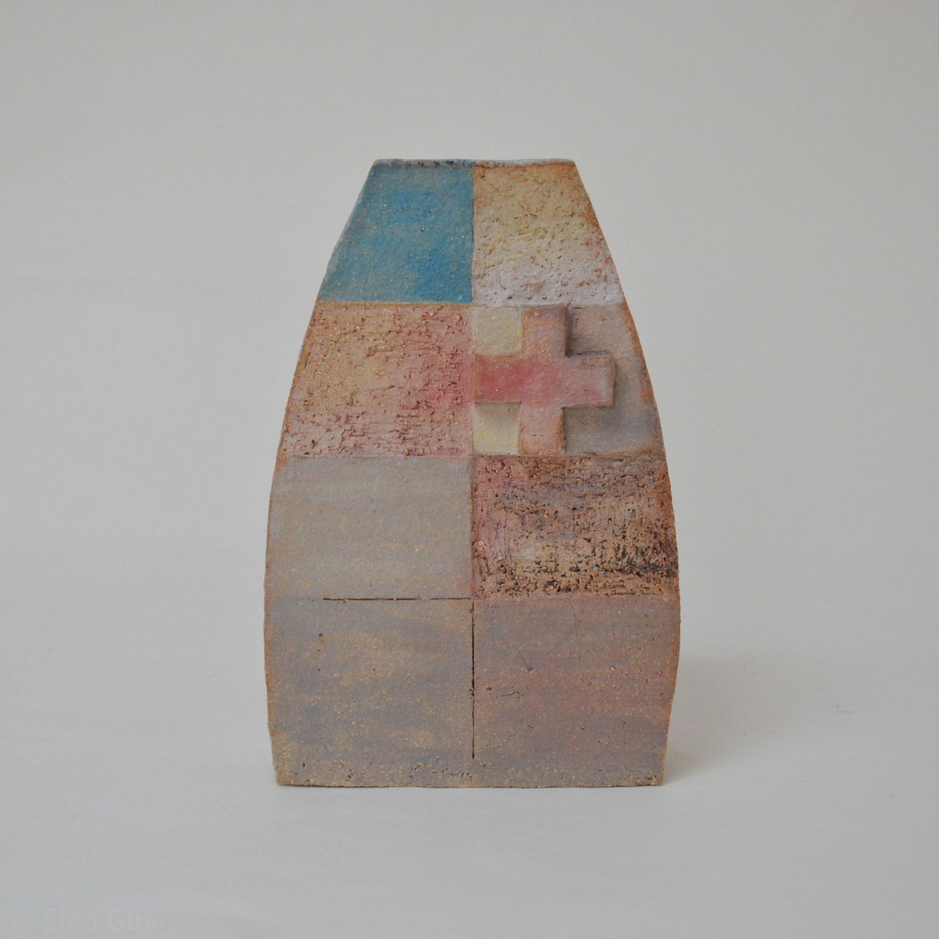 A ceramic sculpture with a trapezoidal top, featuring a grid of textured, muted-toned squares and a small cross relief.