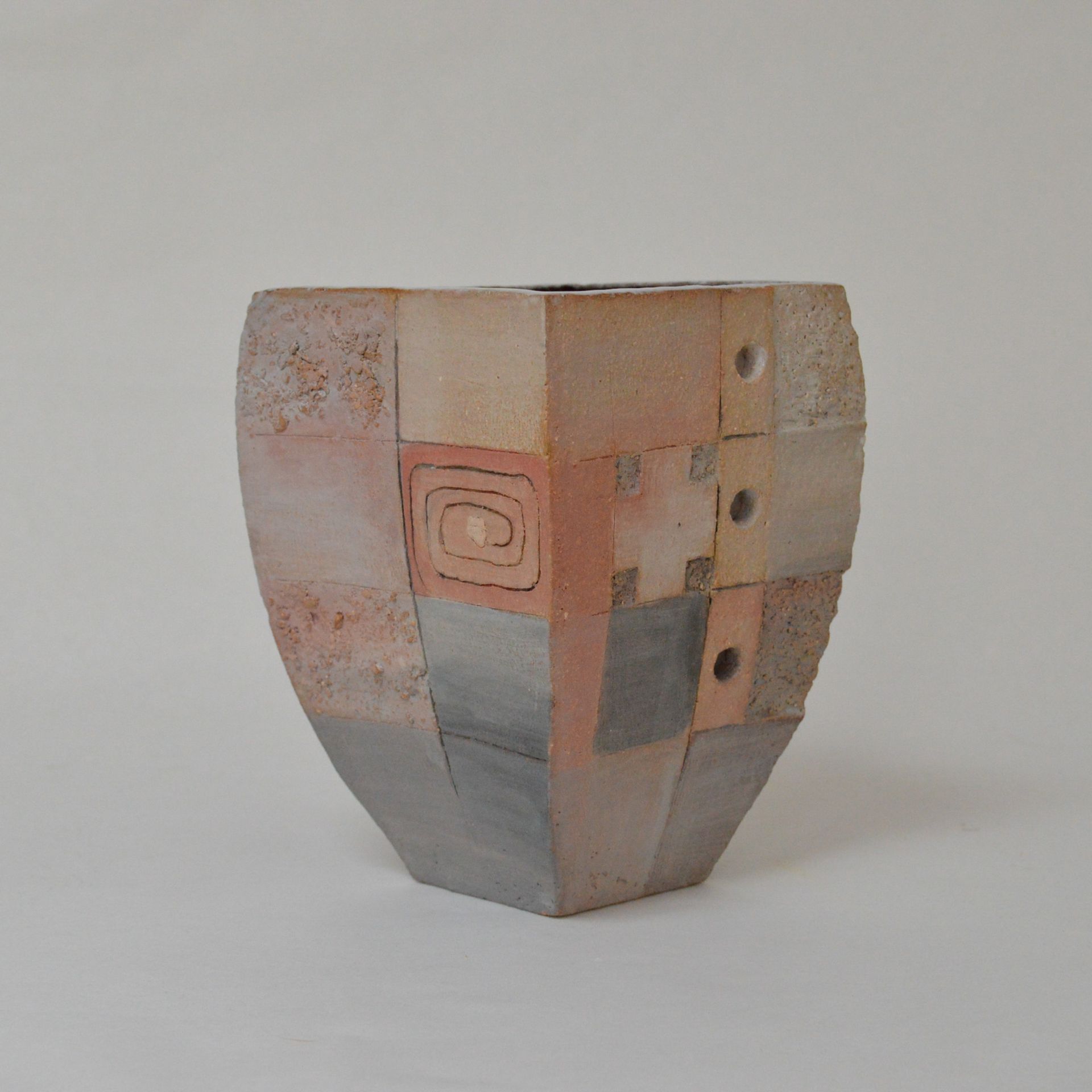 A multi-faceted ceramic vase featuring a patchwork of muted earth tones, a spiral pattern, and three small, aligned holes.