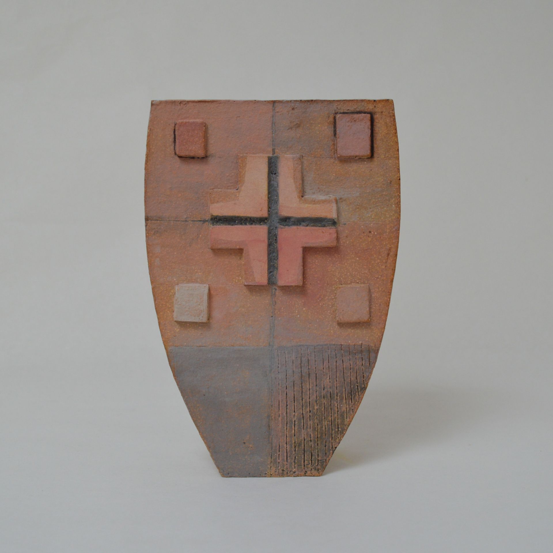 A shield-shaped ceramic sculpture in earth tones, featuring a central cross, four small raised squares, and textured base.