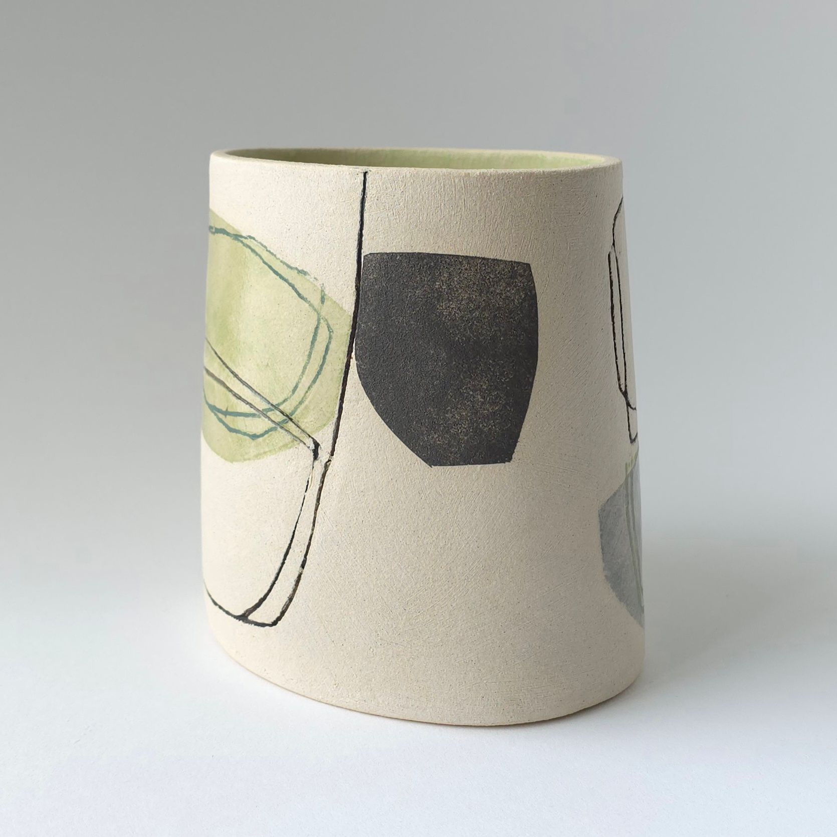 A cylindrical ceramic vase with a speckled beige surface featuring abstract charcoal drawings and a muted green wash.
