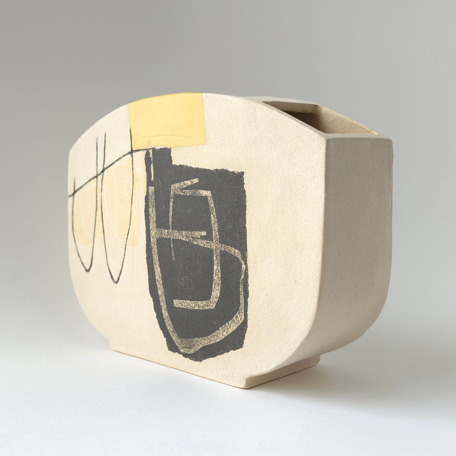 A textured beige ceramic vase with a flat profile, featuring a black abstract geometric design and a yellow accent.
