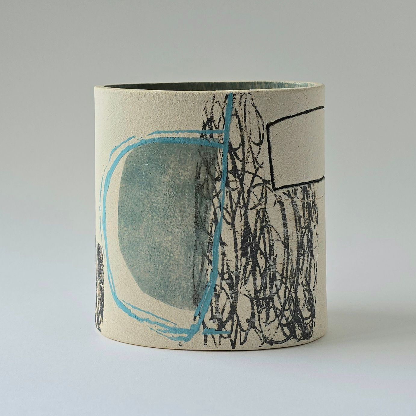 A cylindrical ceramic cup with a speckled beige surface featuring abstract black scribbles and a light blue shape.