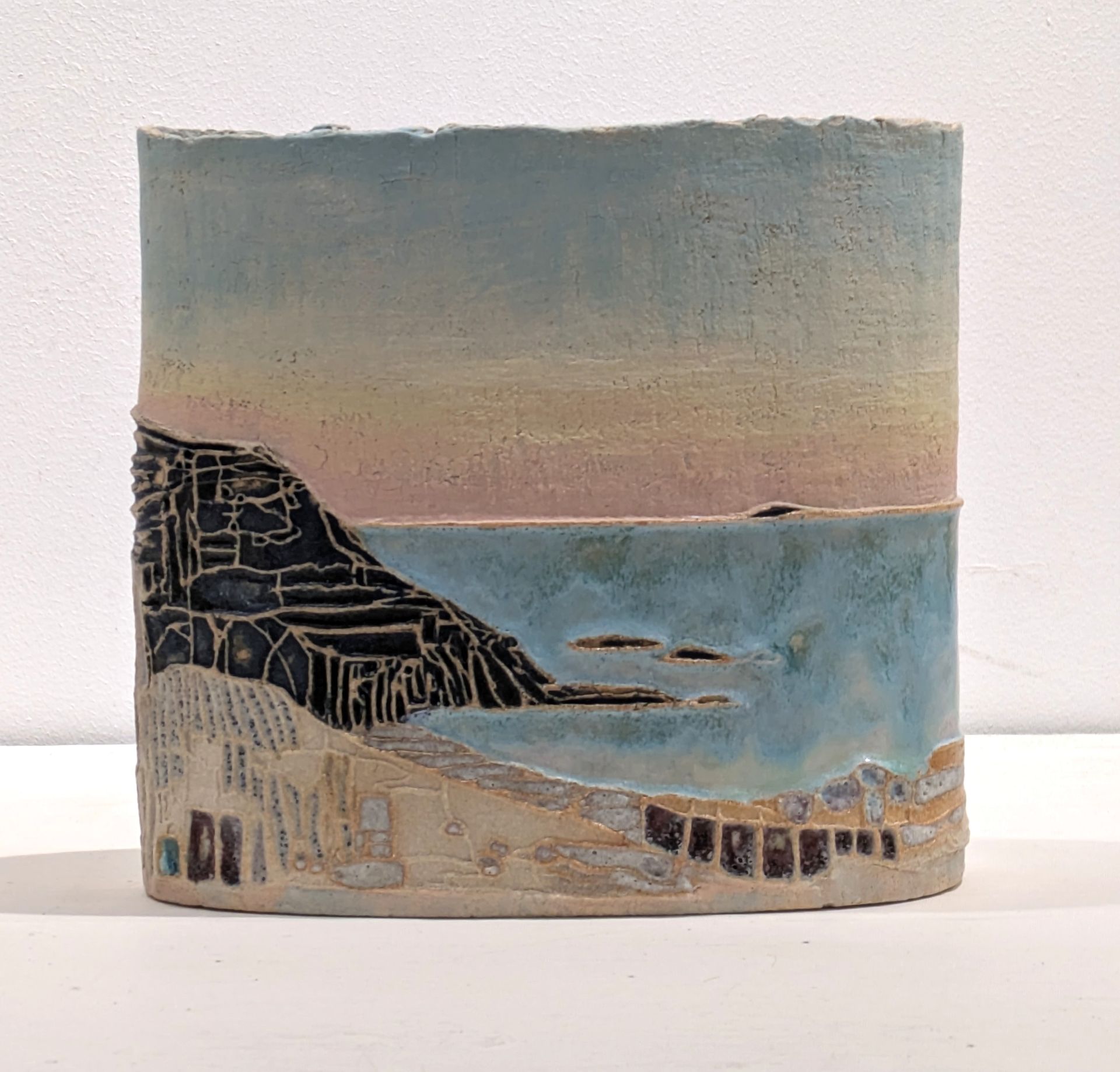 A ceramic vessel featuring a textured, painted coastal scene with a black cliff, blue sea, and sandy shore at dusk.