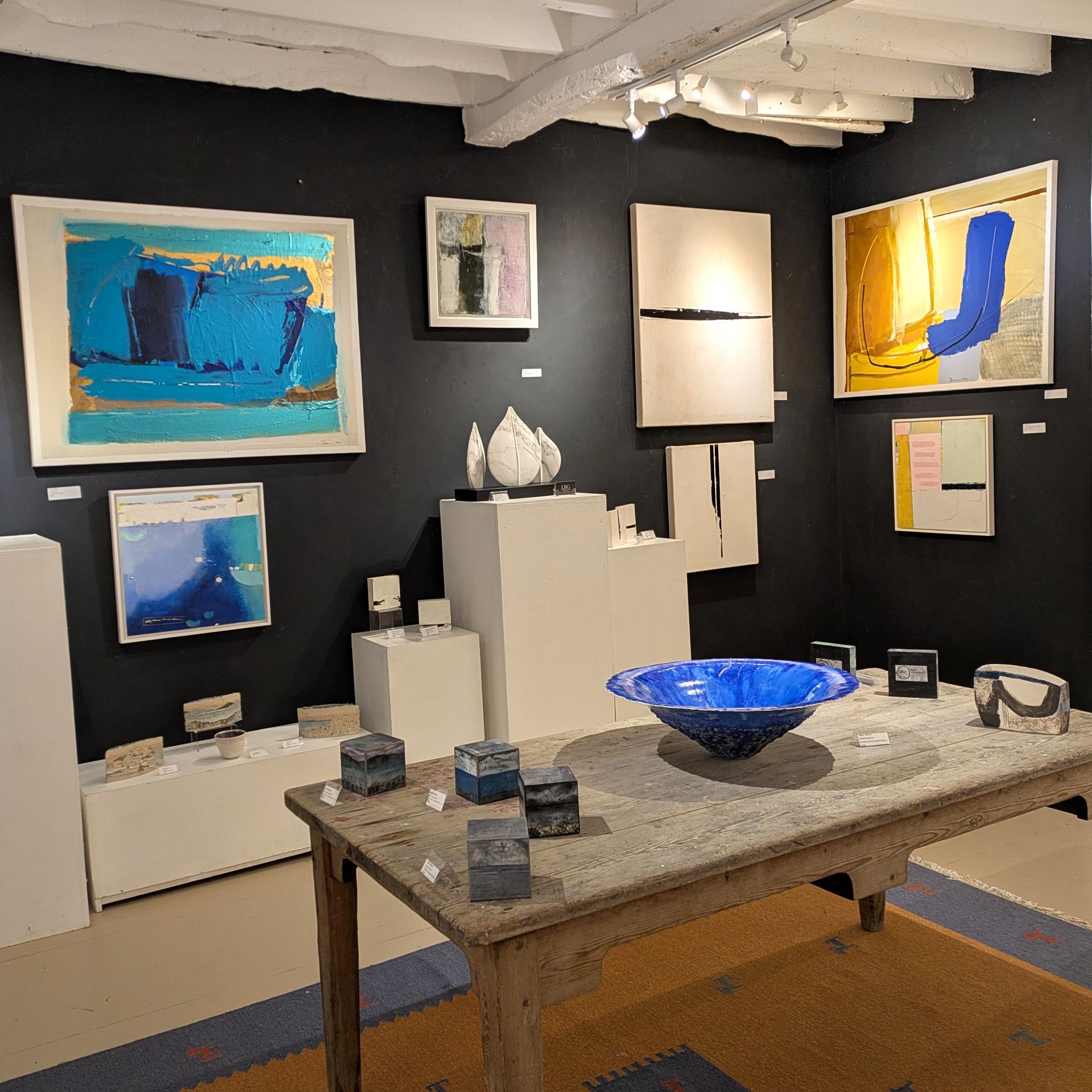 An art gallery interior featuring abstract paintings and ceramic sculptures, with a weathered wooden table in the foreground.