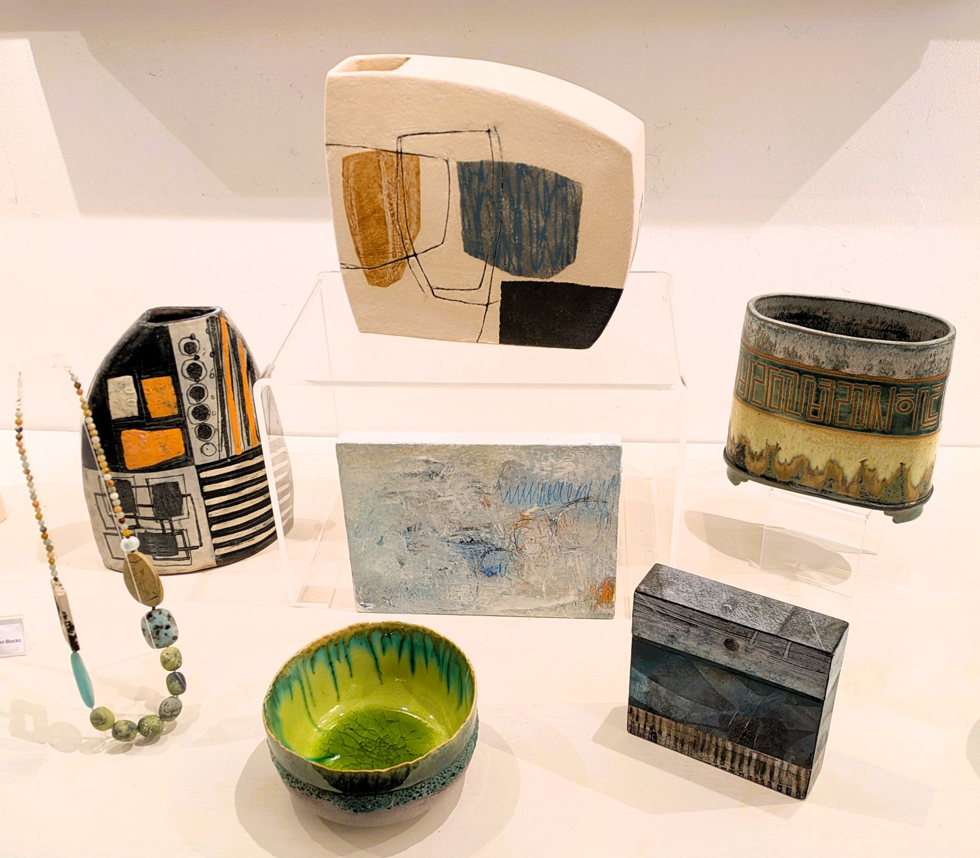 Ceramic art display: vases, bowl, necklace, featuring abstract patterns, neutral and vibrant colors.