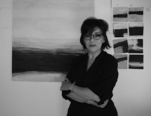 A woman wearing glasses is standing in front of a painting on a wall.