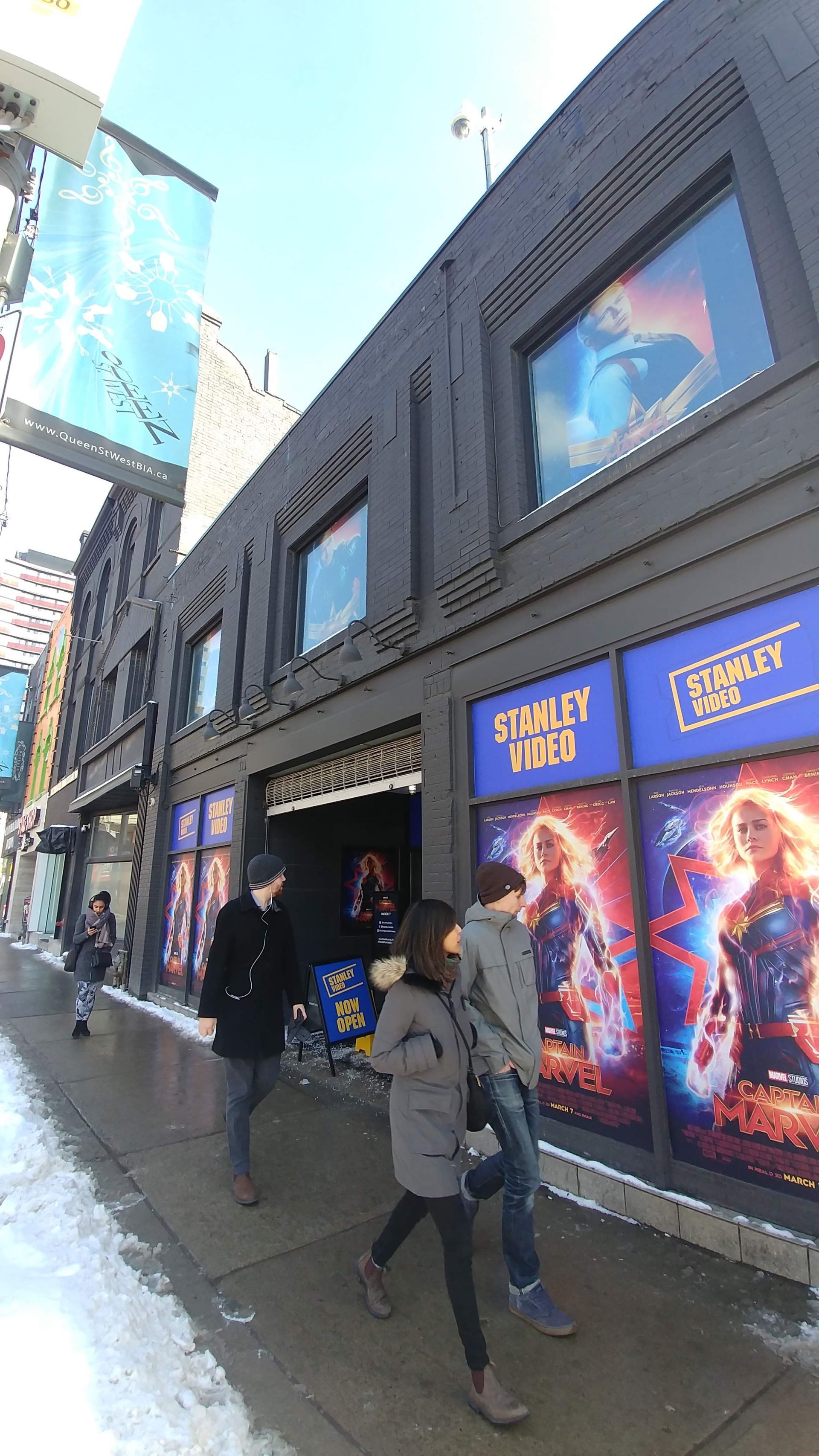 A group of people are walking down a sidewalk in front of a building with captain marvel posters on the windows.