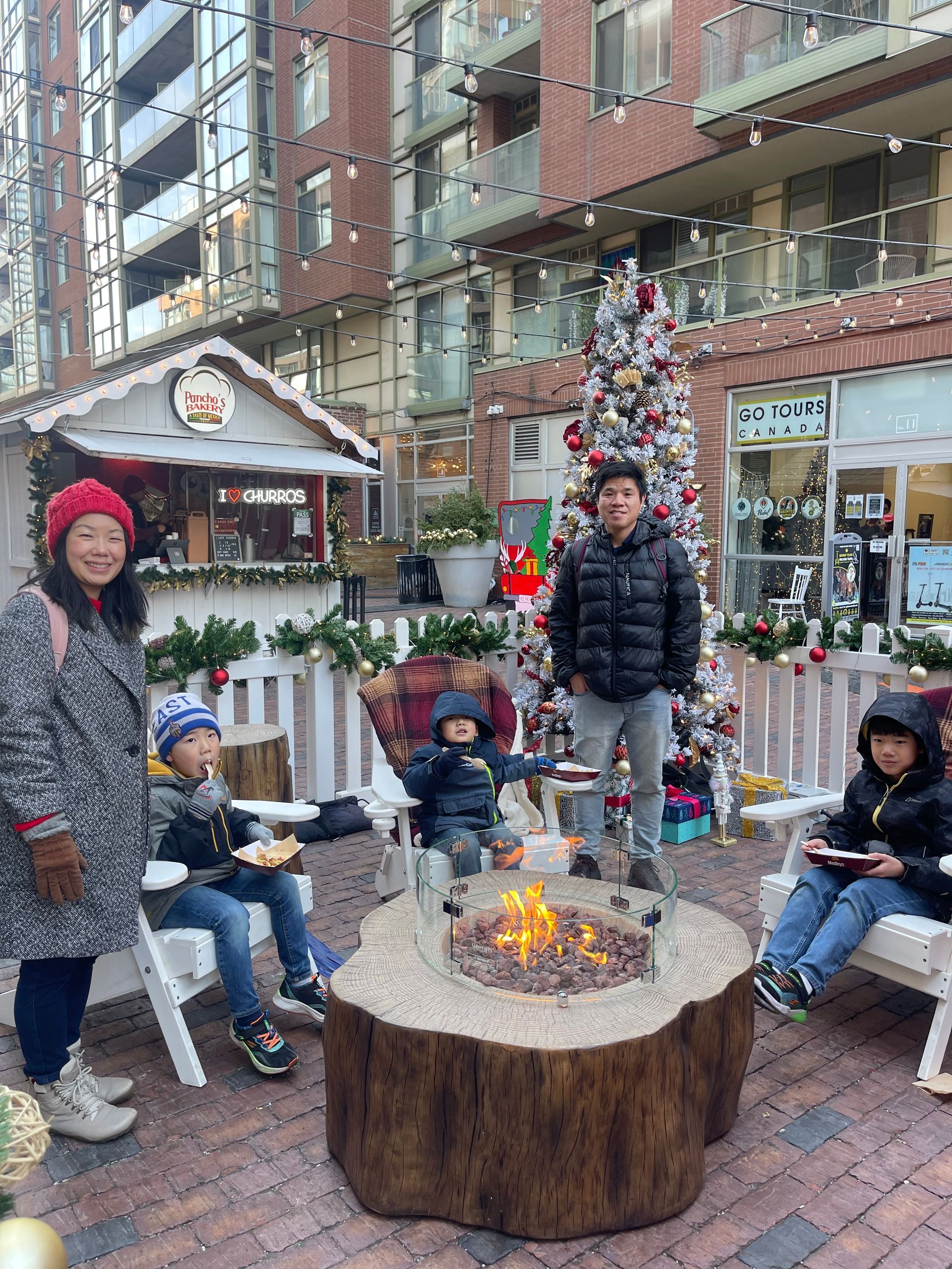 A family is sitting around a fire pit in front of a christmas tree.