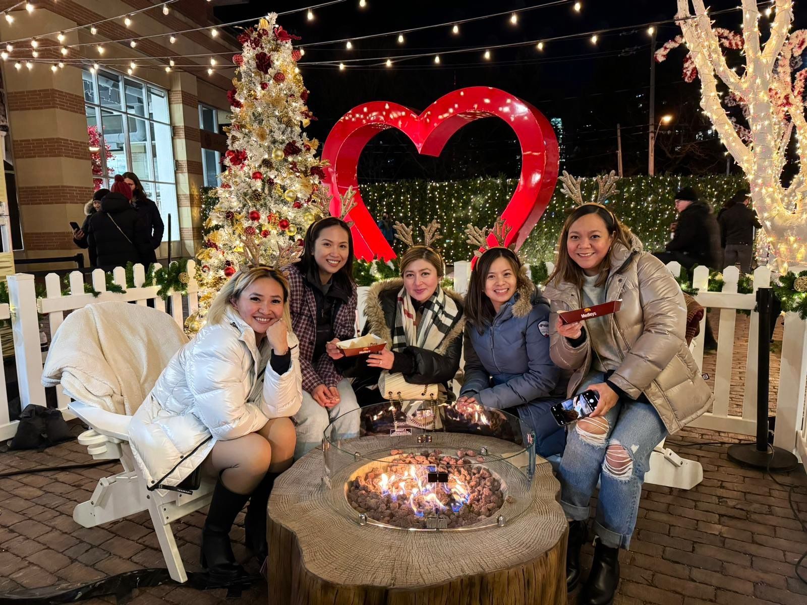 A group of women are sitting around a fire pit in front of a christmas tree.