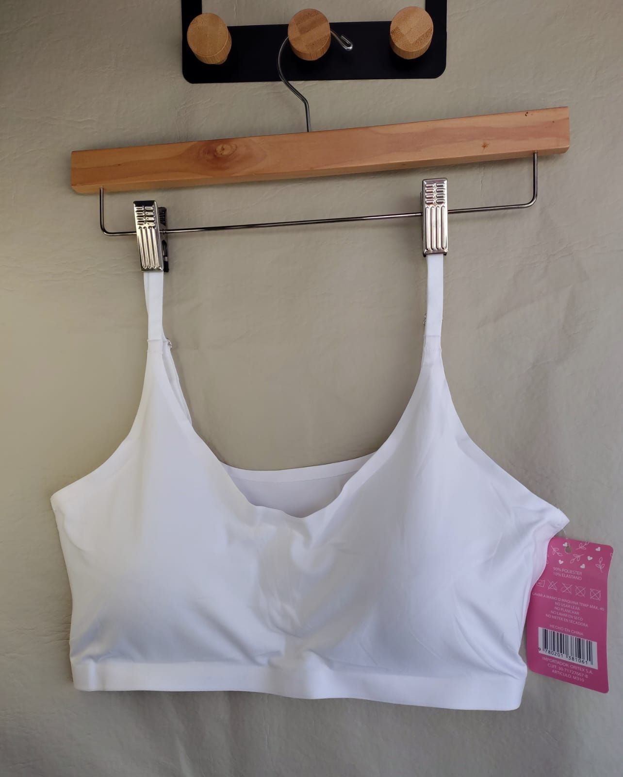 Sujetador blanco con tirantes finos colgado en una percha de madera, con etiqueta rosa.