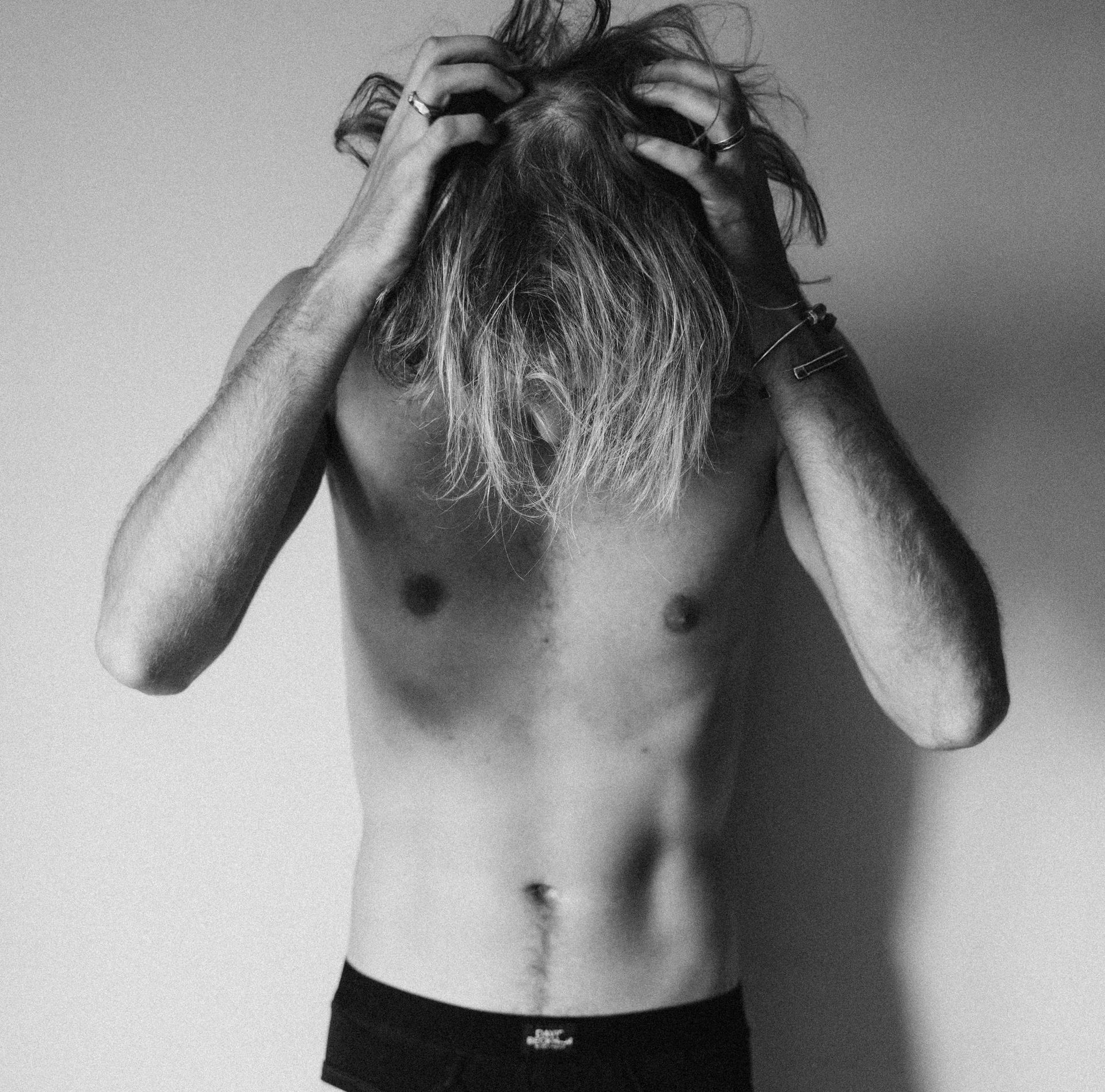 Hombre con cabello largo rubio, torso desnudo, manos en el cabello, vistiendo ropa interior negra.