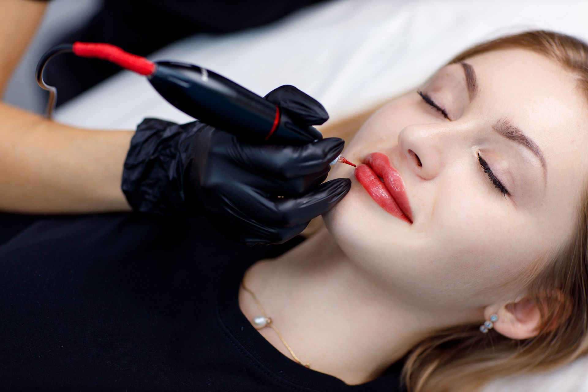 Woman getting lip blushing procedure. Close-up shows gloved hand holding tattoo pen applying pigment to lips.