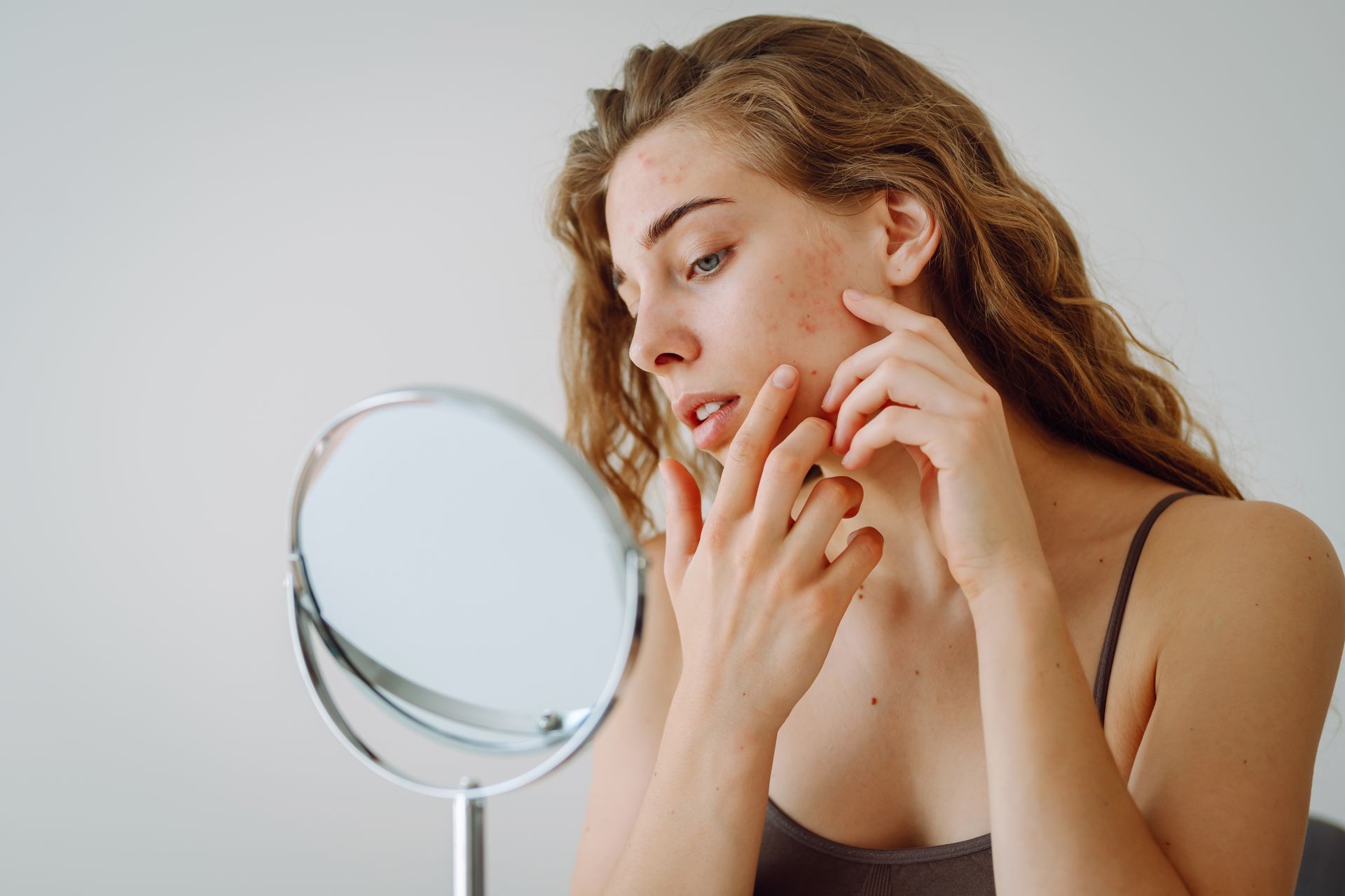 Woman with acne looks in a mirror, touching her cheek. Indoors, neutral background.
