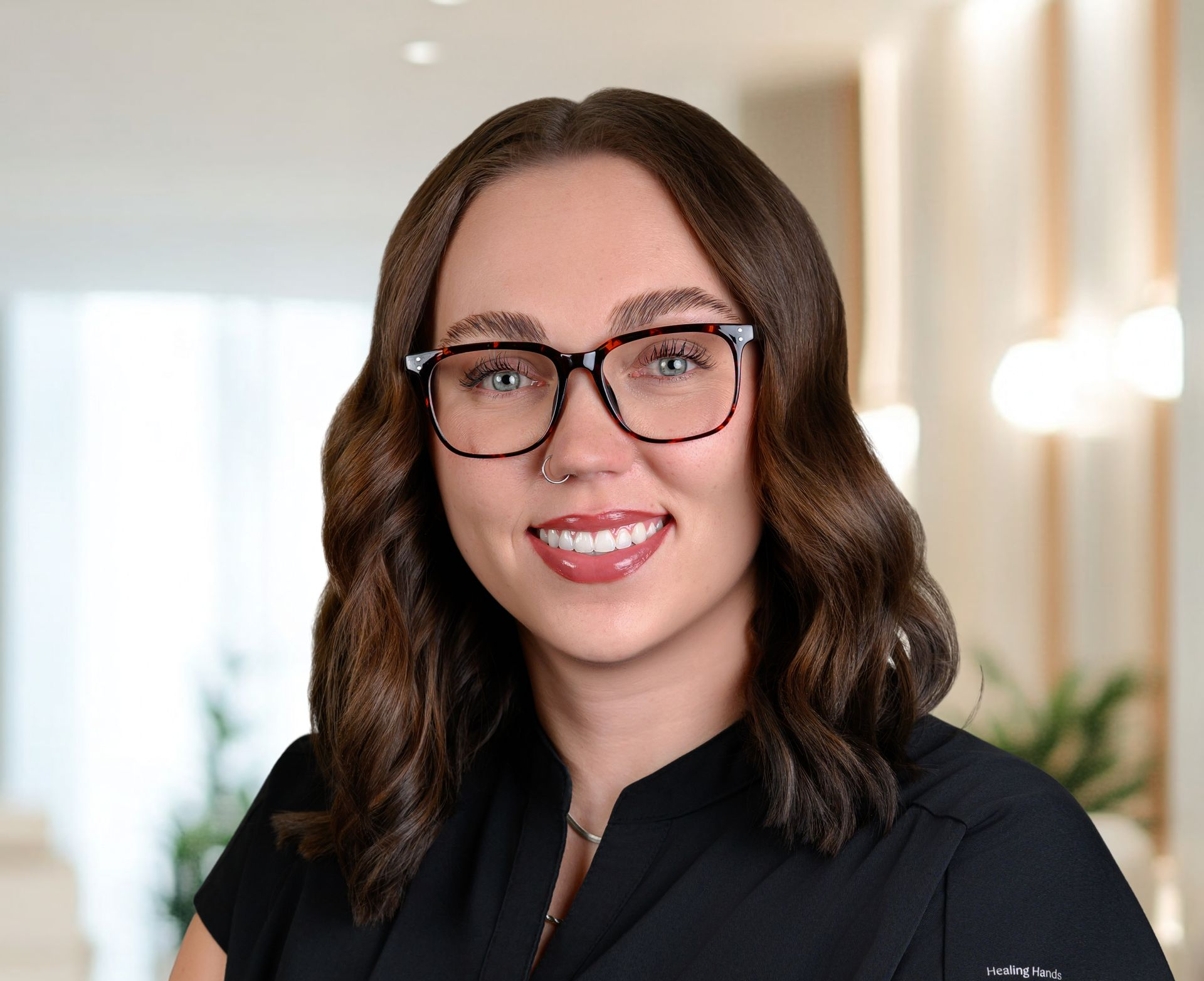 Woman with brown hair, glasses, and a nose ring smiles. She is wearing a black shirt in a bright office setting.