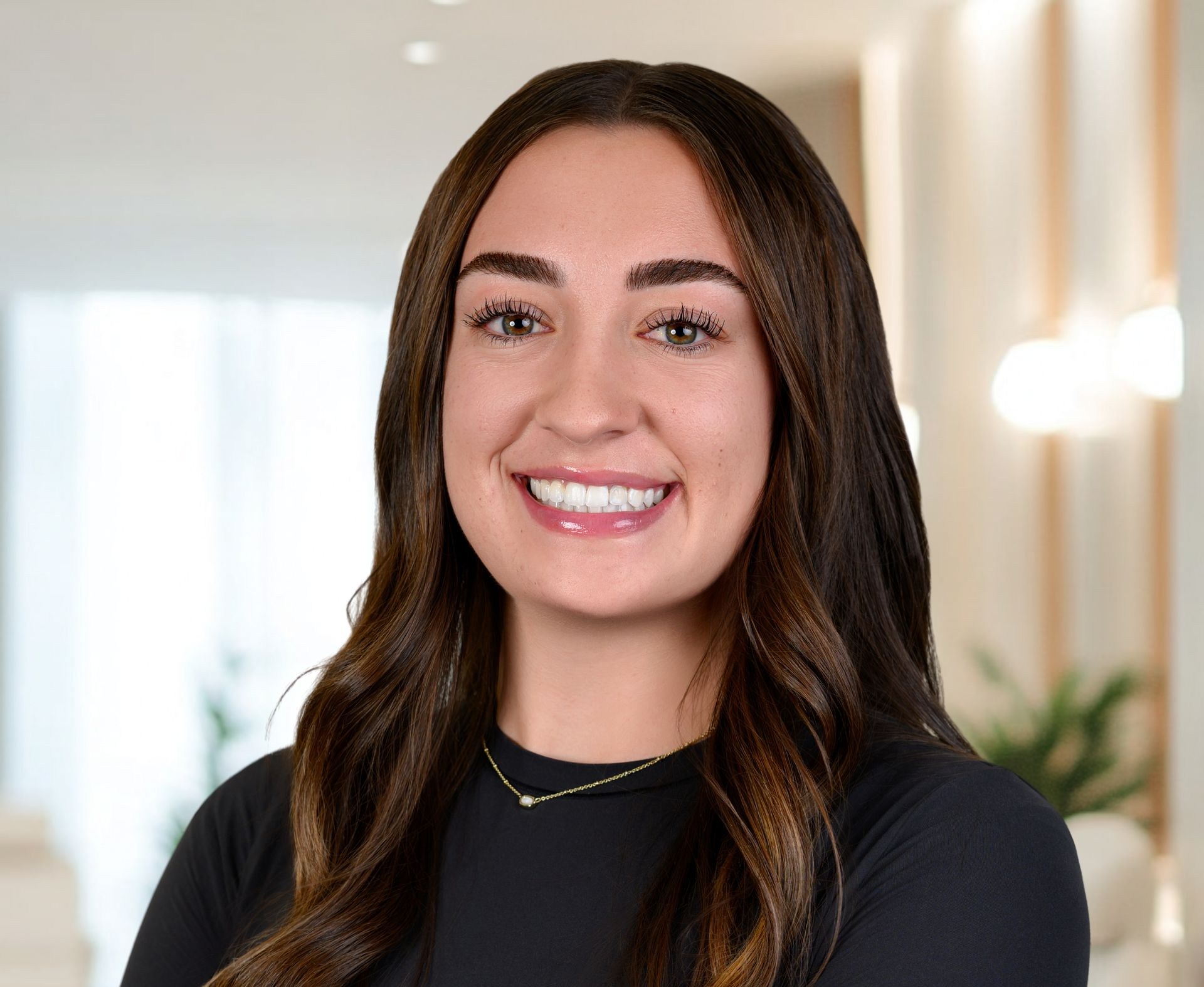 Woman smiling, brown hair, black top, gold necklace. Blurred office background.