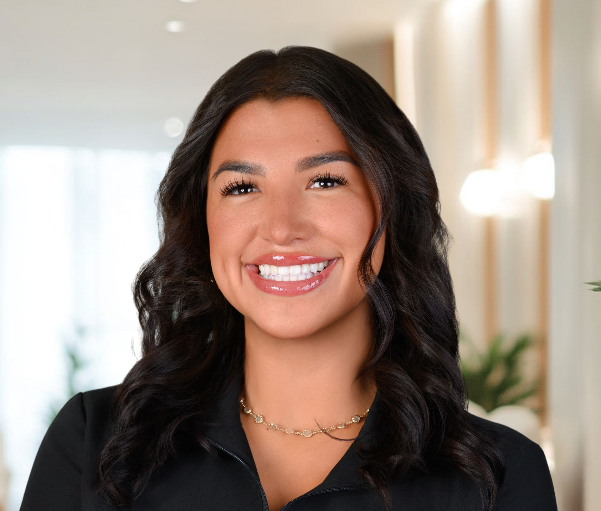 Woman with dark hair, smiling broadly, wearing a black blazer and gold necklace.