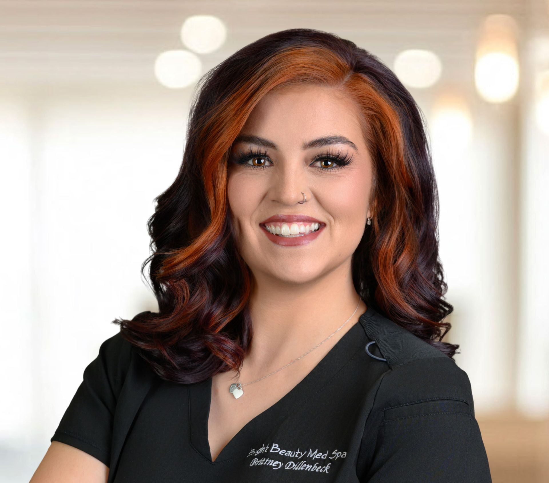 Woman with dark and red hair, smiling, wearing a black top, small necklace. Blurred background.