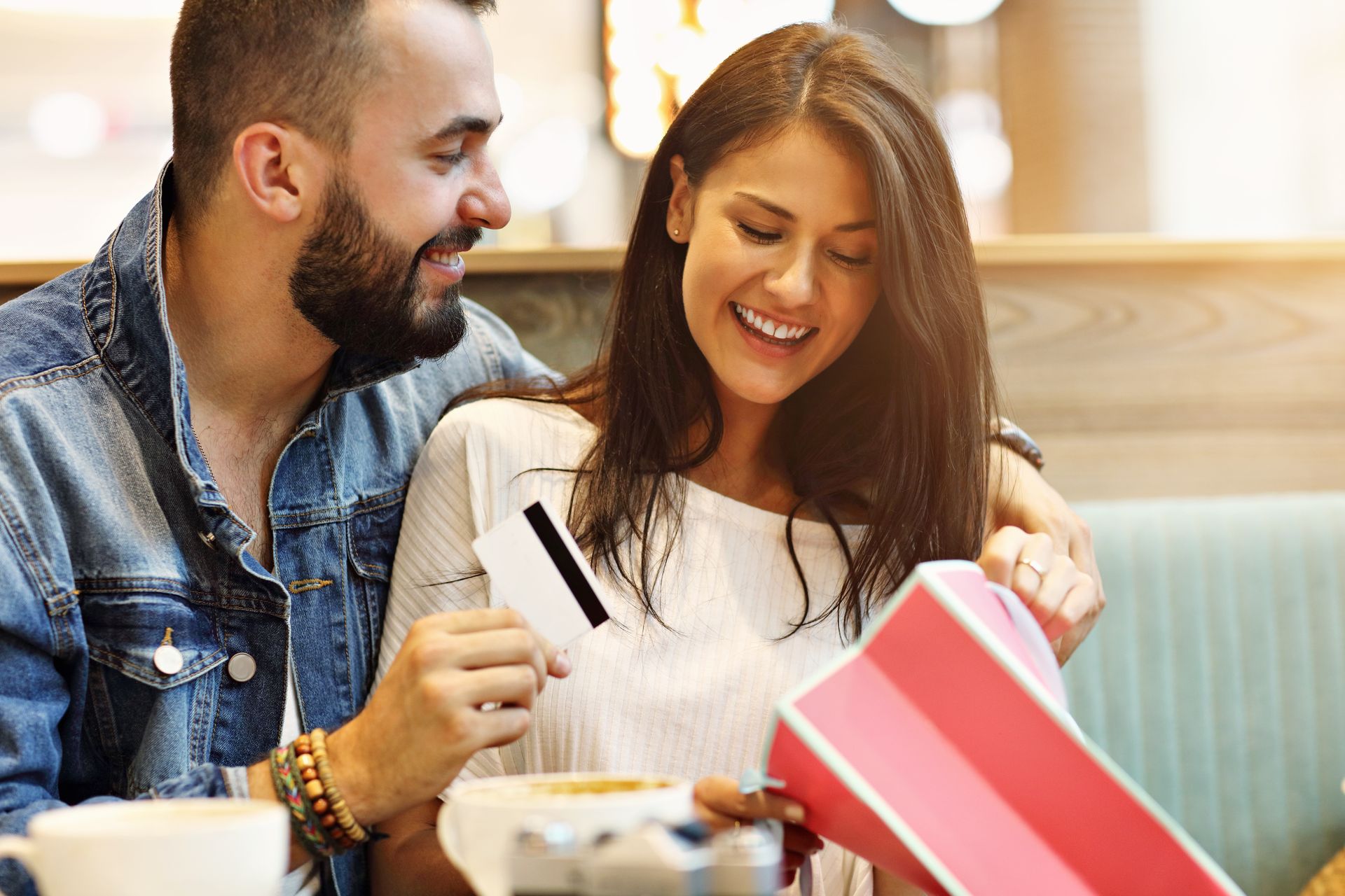 Couple at a cafe: man looks on as woman smiles, holding a credit card and gift box, possibly shopping.