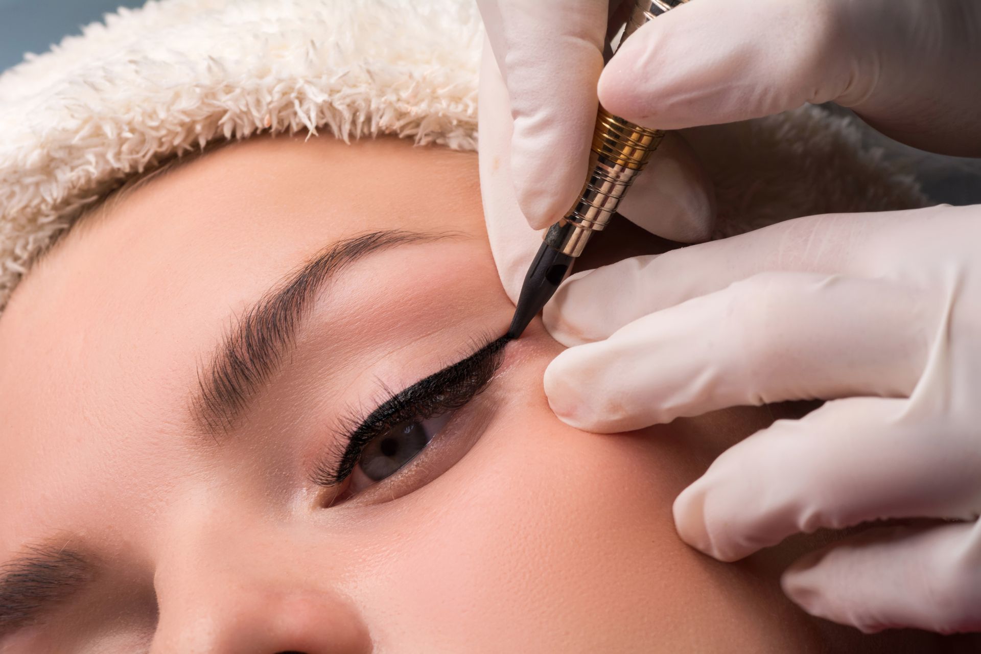 Eyeliner being tattooed on a person’s upper eyelid by a gloved hand.