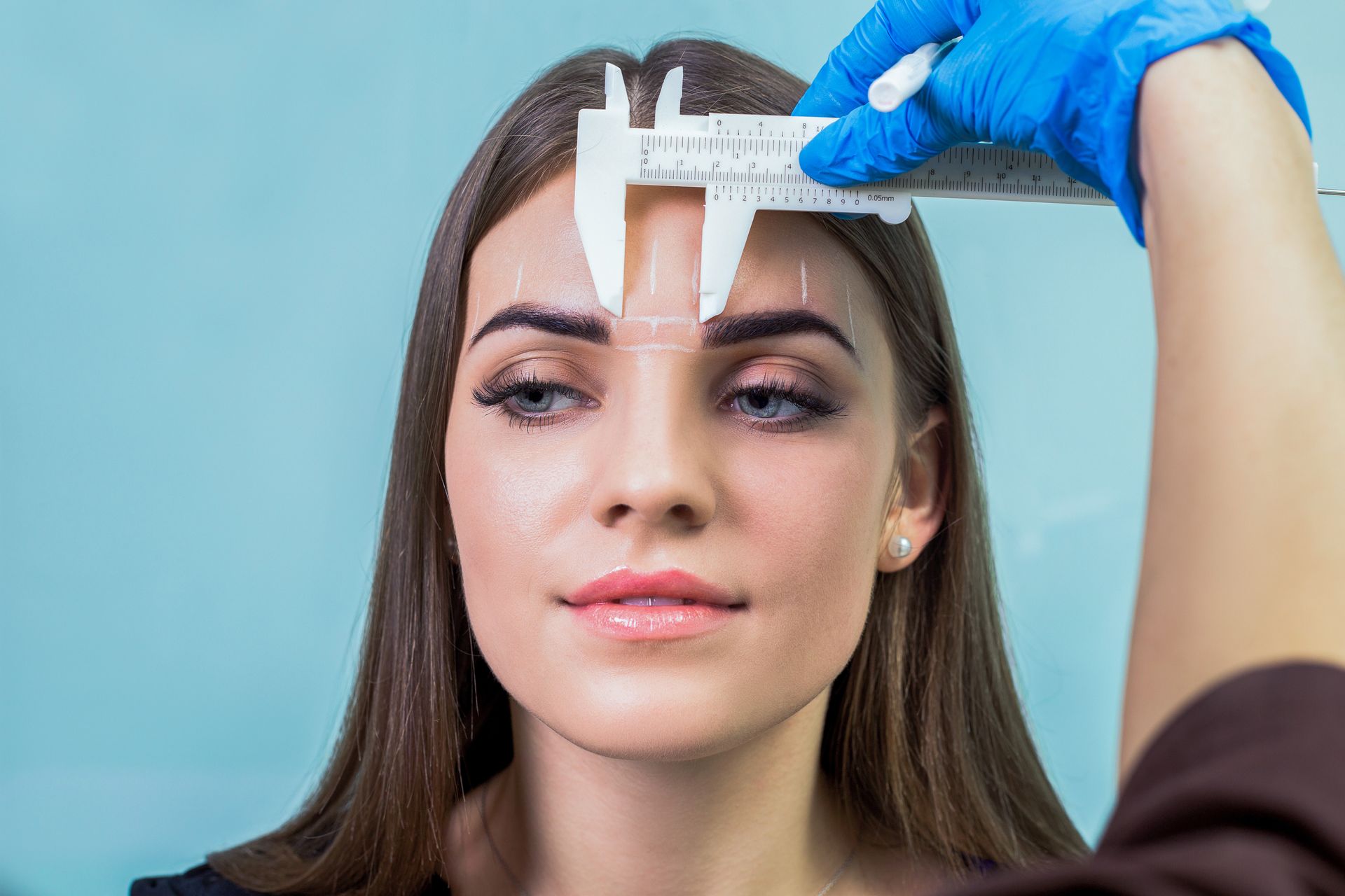 Woman's face with eyebrows marked; technician in blue gloves measures brows with calipers, light blue background.