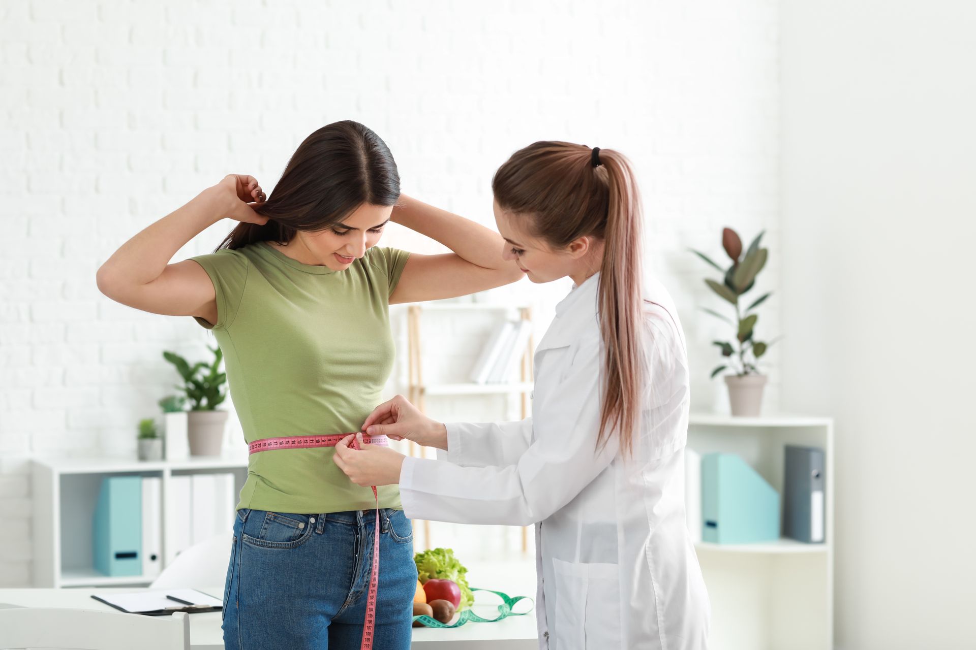 Woman having waist measured by a person in a lab coat in an office with a white background.