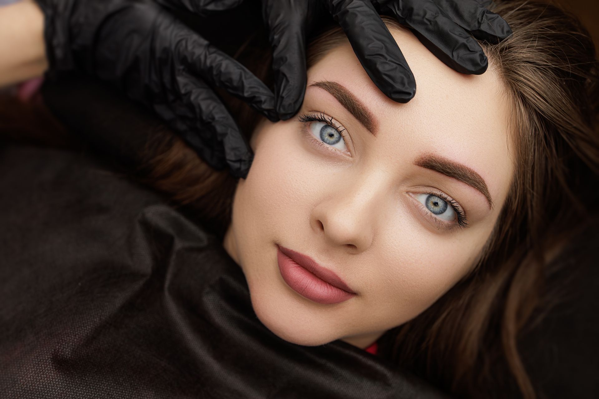 Woman with new eyebrow tattoo, gloved hand checking shape. Brown eyebrows, blue eyes, dark setting.