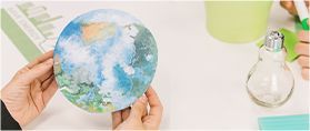 Hands holding a paper globe; a light bulb, and a green object sit on a white surface.