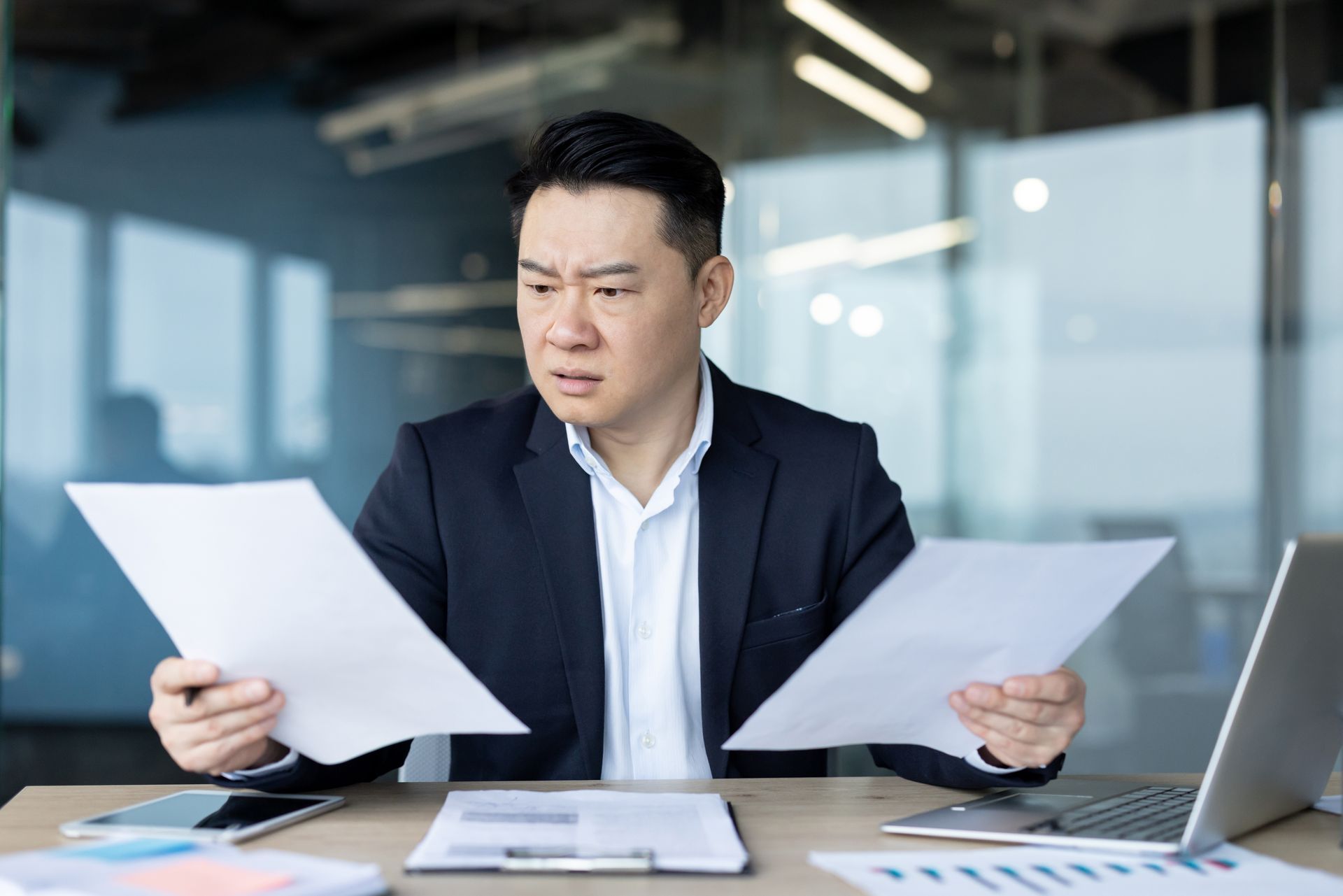 A businessman sits in an office at a desk and looks disappointedly at documents he is holding.