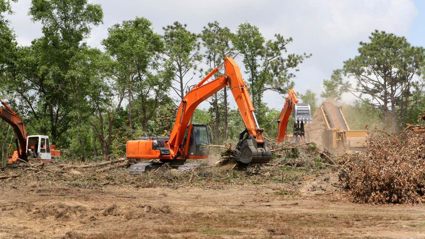 Orange excavators clearing brush at a construction site under a cloudy sky