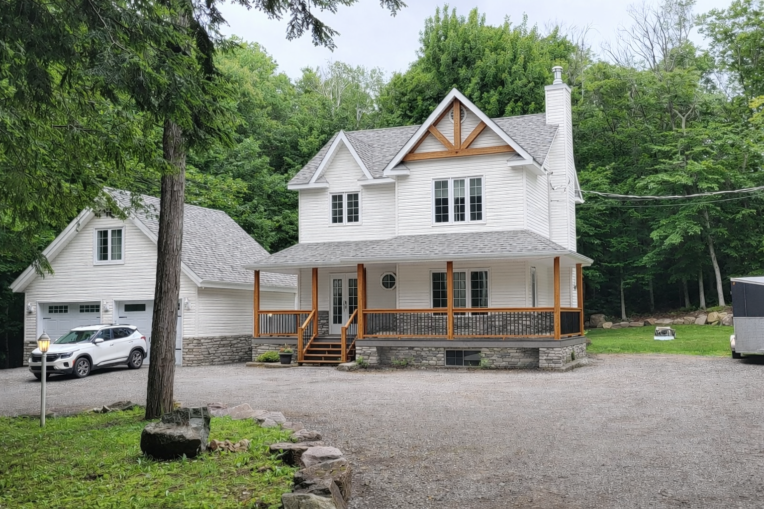 Une maison blanche de deux étages avec une véranda en bois qui l'entoure, un soubassement en pierre et un garage attenant, située dans une zone boisée.