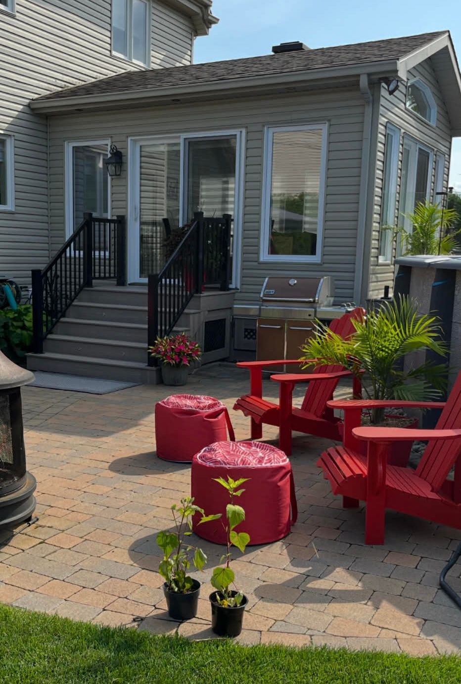 Une terrasse arrière avec des pavés de brique claire, deux chaises Adirondack rouges, des poufs rouges et des plantes en pot près de l'escalier.