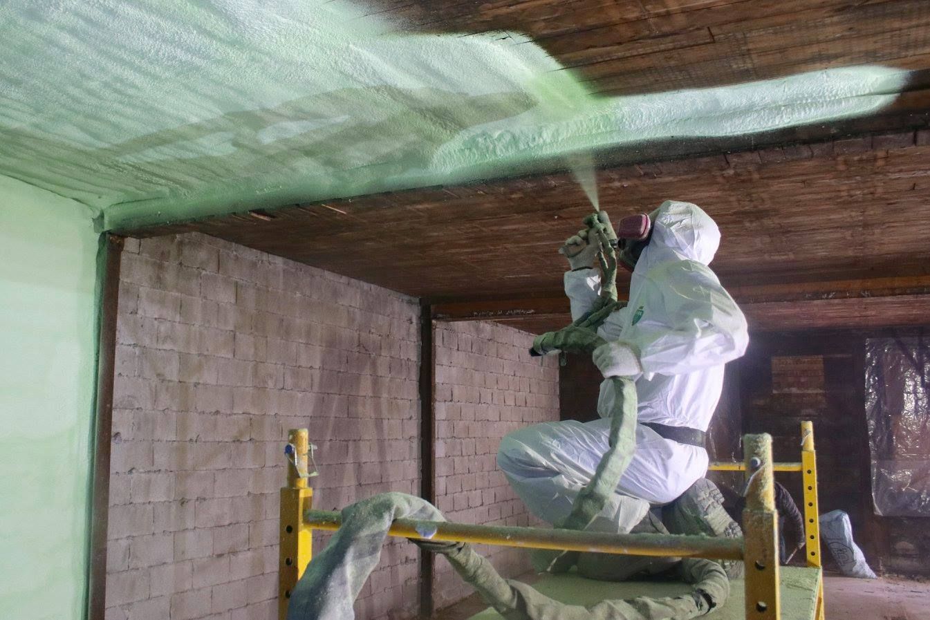 Une personne en combinaison de protection pulvérise de la mousse isolante sur un plafond en bois tout en se tenant sur un échafaudage jaune.