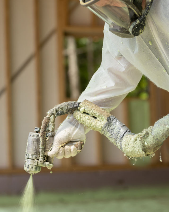 Un ouvrier en tenue de protection utilise un pistolet à peinture professionnel pour appliquer de l'isolant en mousse verte sur une ossature murale.