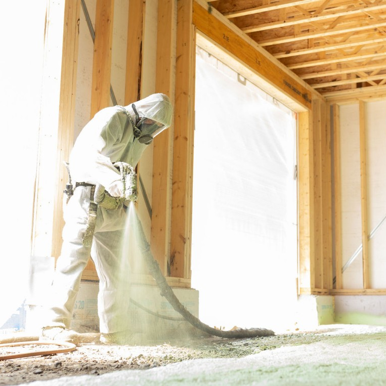 Un ouvrier vêtu d'une combinaison de protection blanche et d'un masque respiratoire pulvérise de l'isolant en mousse sur le sol d'un bâtiment à ossature bois.