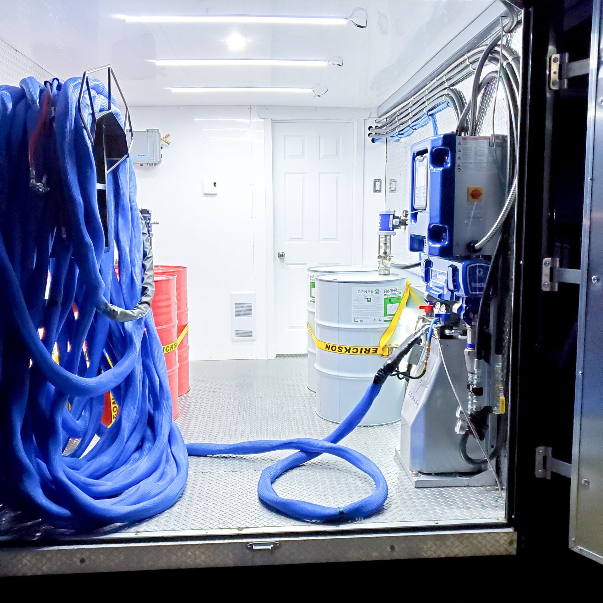 À l'intérieur d'une remorque utilitaire blanche, un tuyau bleu est enroulé à côté de fûts industriels et d'un système de pompage hydraulique.