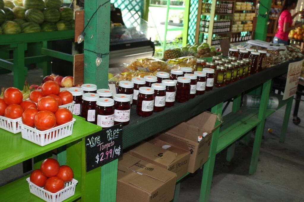 Produce stand with tomatoes, jams, and other goods on green tables.