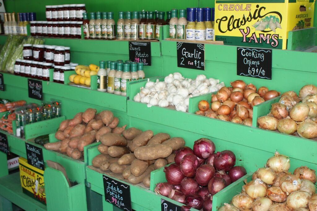 Produce display in a market with jars, bottles, onions, potatoes, and garlic on green shelves.