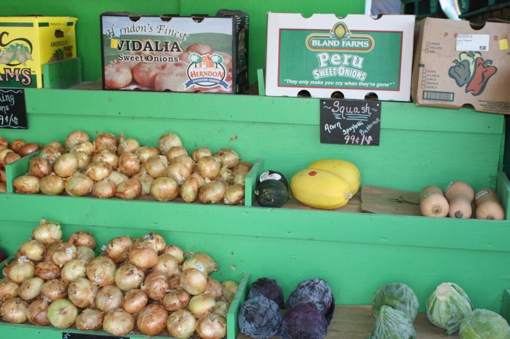 Produce display: onions, squash, and plums on green shelves, with boxes overhead.