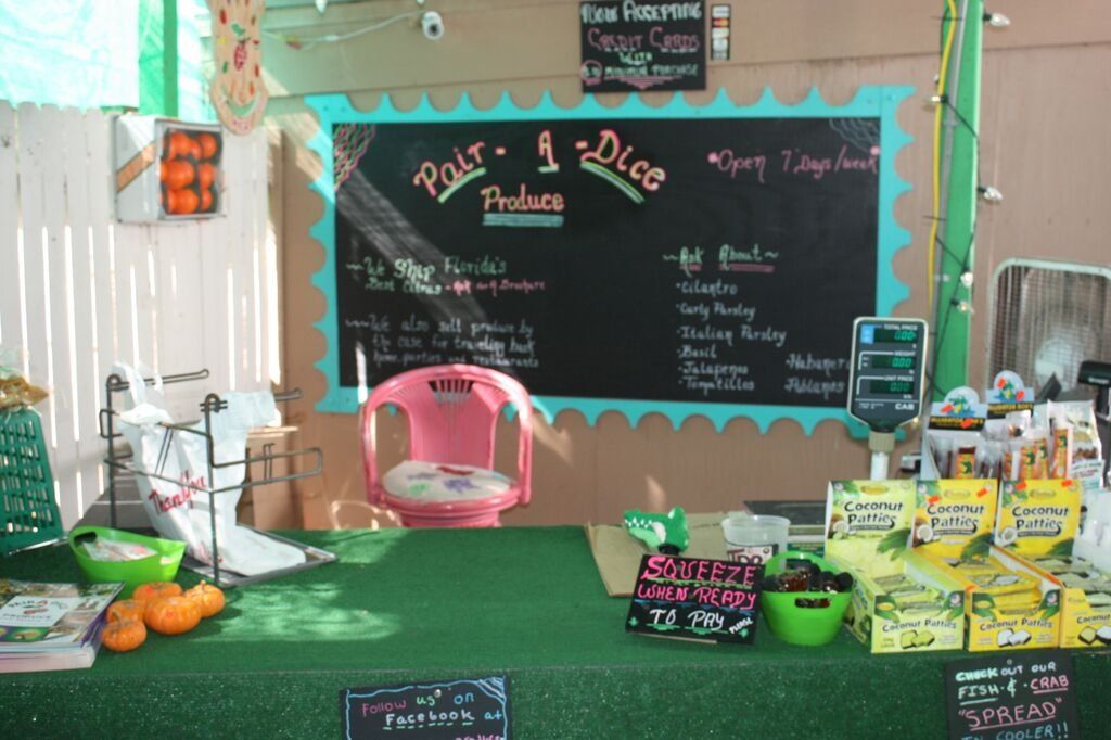 Produce stand with a chalkboard menu and a pink chair. Green produce, orange squash, and packages are displayed.