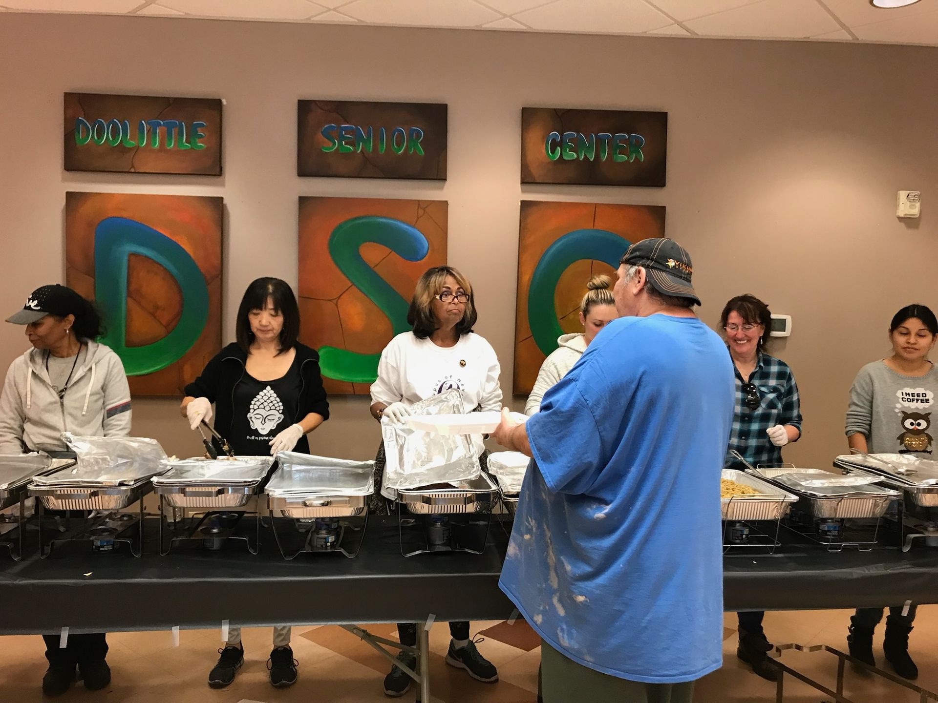 People serving food at the Dexter Senior Center. Individuals in front of serving trays, handing out meals.