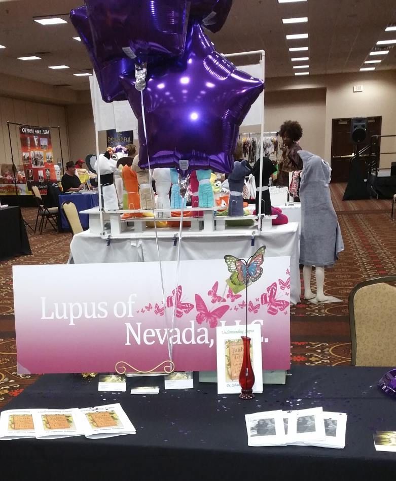 Lupus of Nevada booth at an event with purple balloons and a person browsing. Black table with brochures.